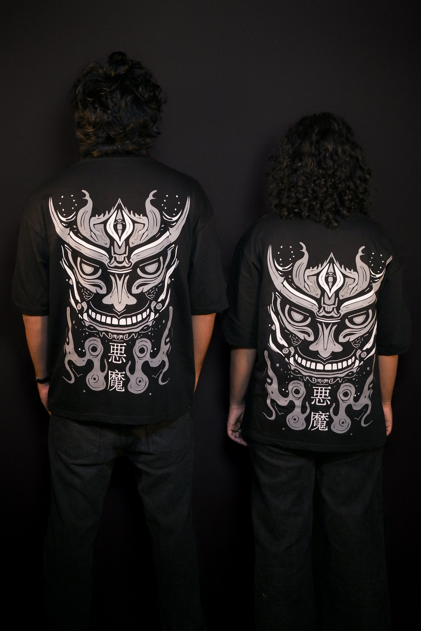 Two people wearing black t-shirts with white graphic designs on the back against a dark background.