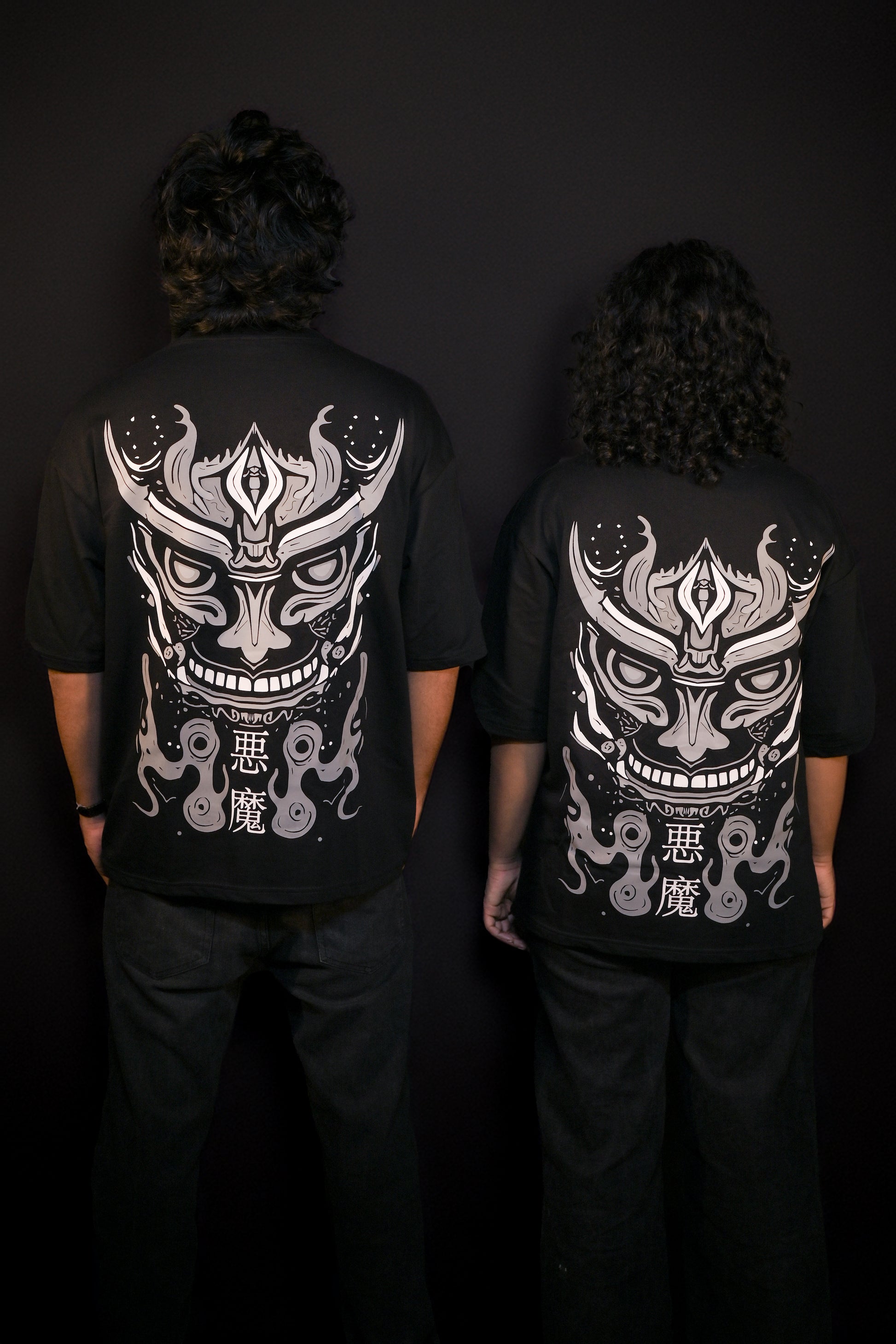 Two people wearing black t-shirts with white graphic designs on the back against a dark background.
